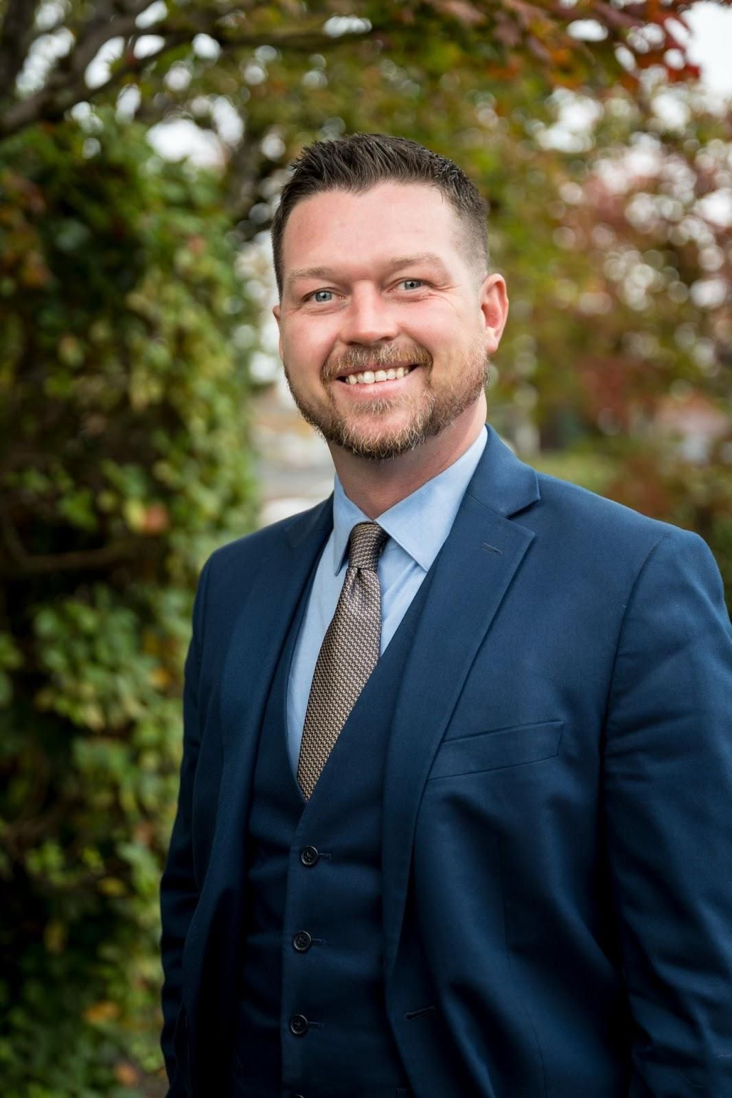Man in a blue suit smiles outdoors, leaning against green foliage.