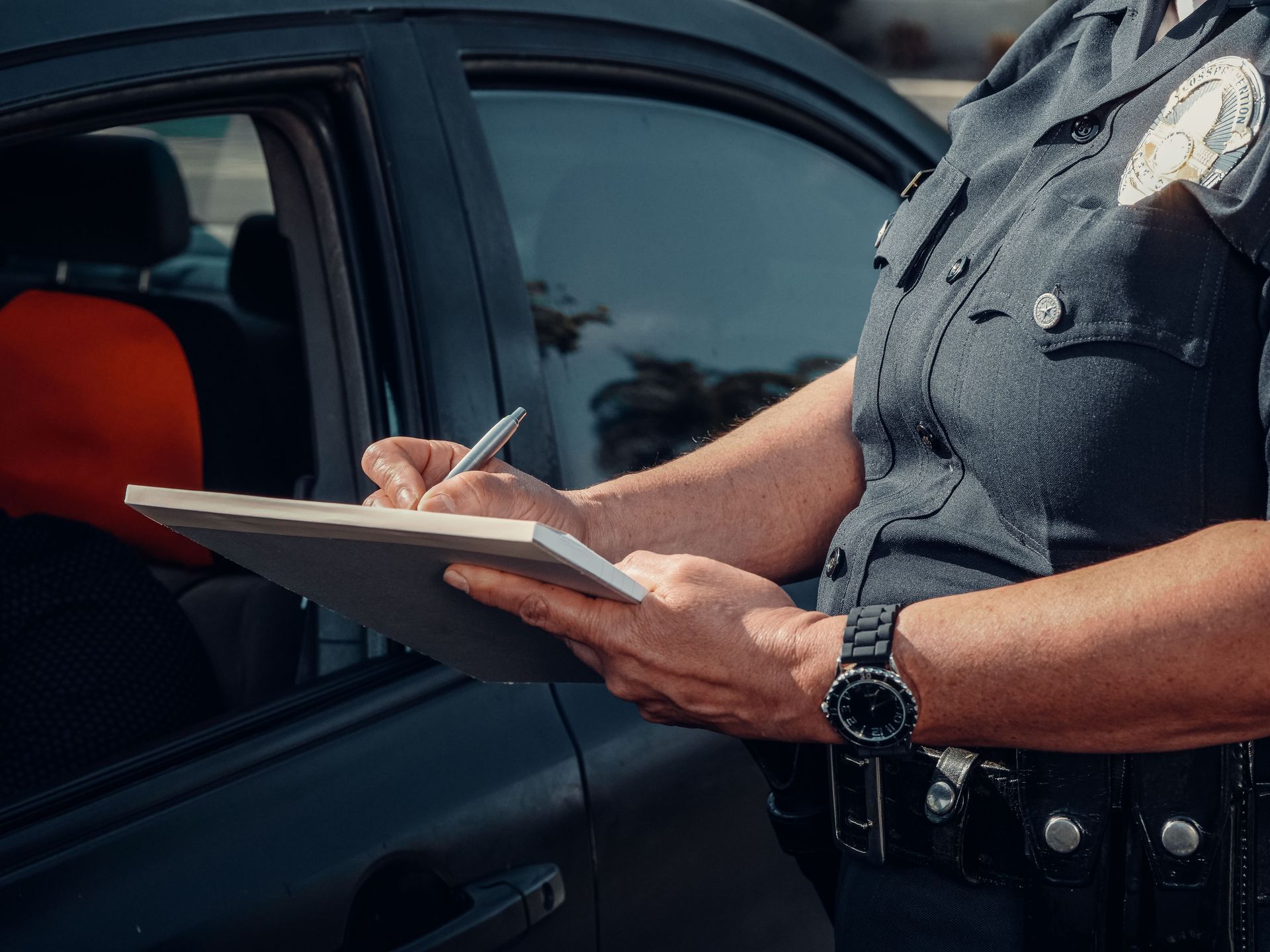 A police officer writing on a pad standing next to a car with the window rolled down.