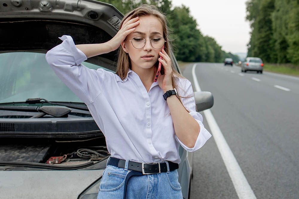 Woman with a broken-down car on the side of a highway, holding her head and talking on a phone.