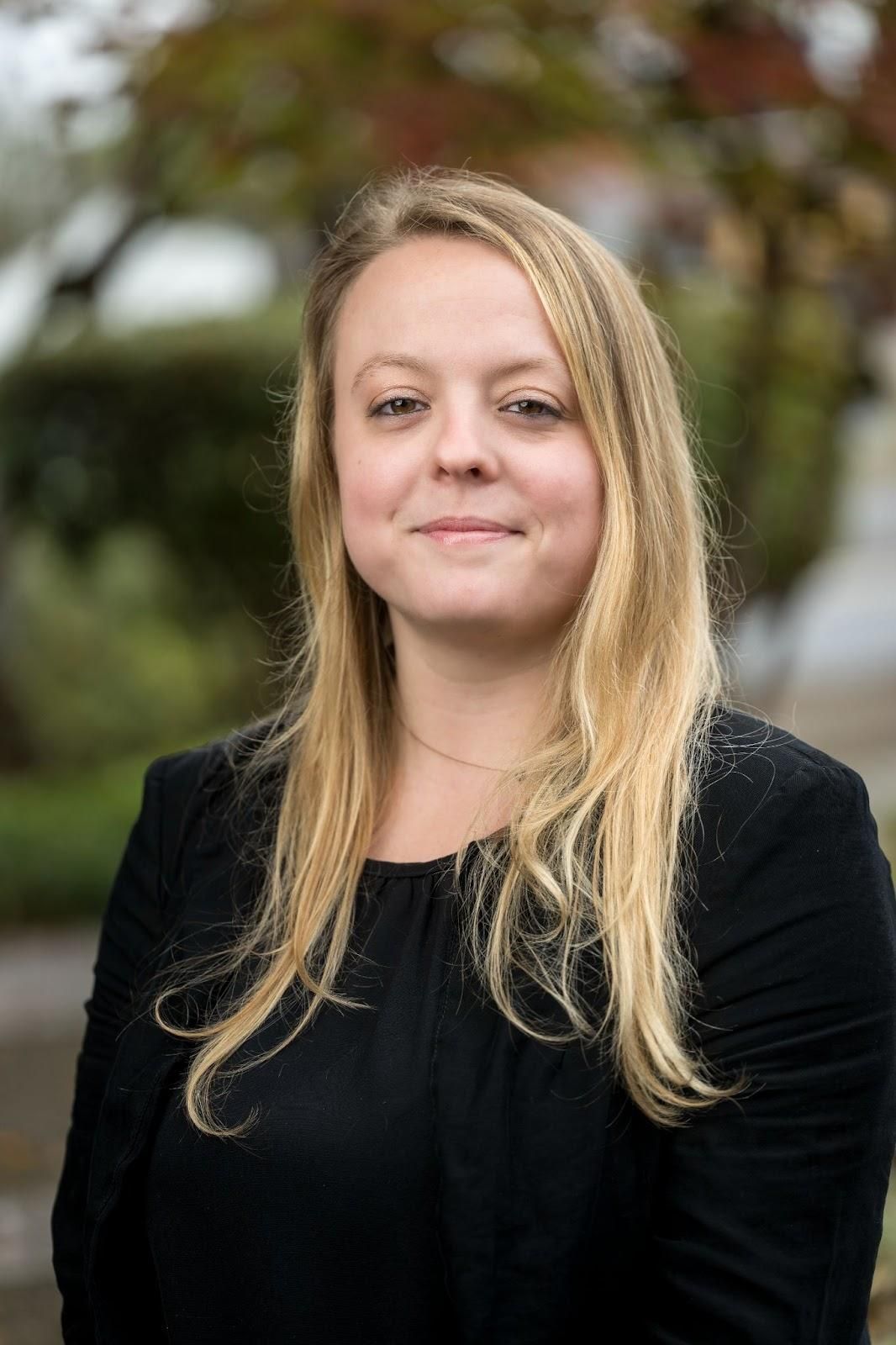 Blonde woman in black blazer smiles, outdoors.