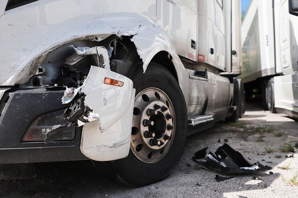 Damaged white semi-truck after a collision; front fender torn and debris on the ground.