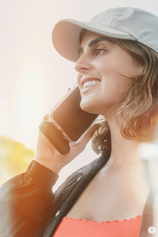 Woman smiling while talking on a smartphone outdoors in sunlight, wearing a white cap and coral top