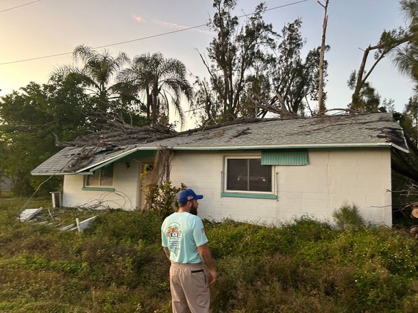 A man is standing in front of a house with a roof that has been damaged by a storm.