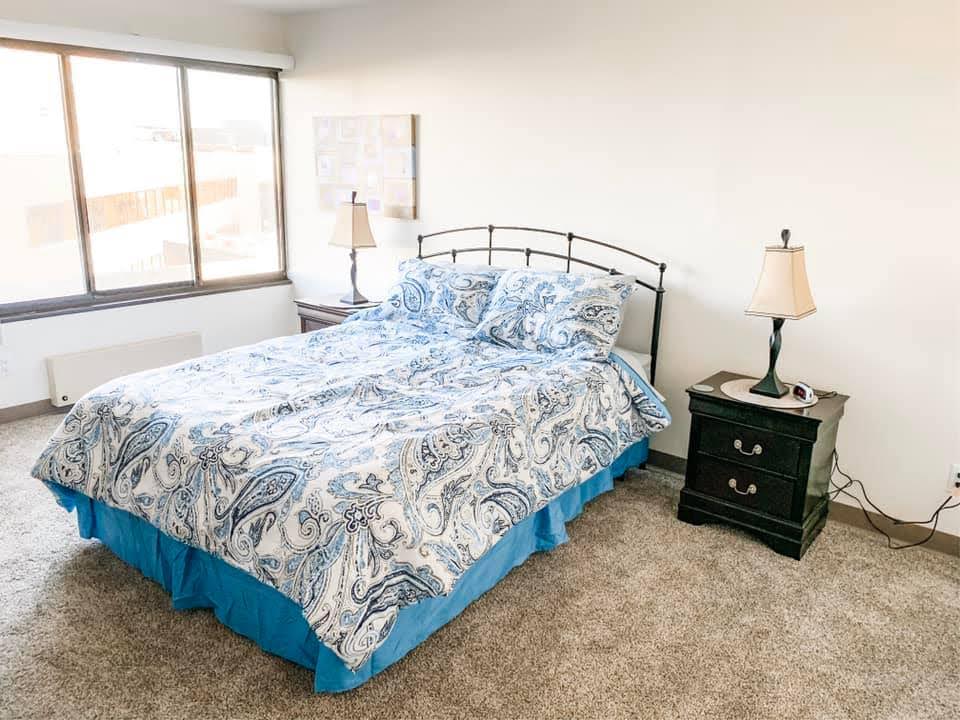 Interior view of apartment bedroom area example with light gray carpet, bed with blue floral comforter, dark wood night stand with lamp.