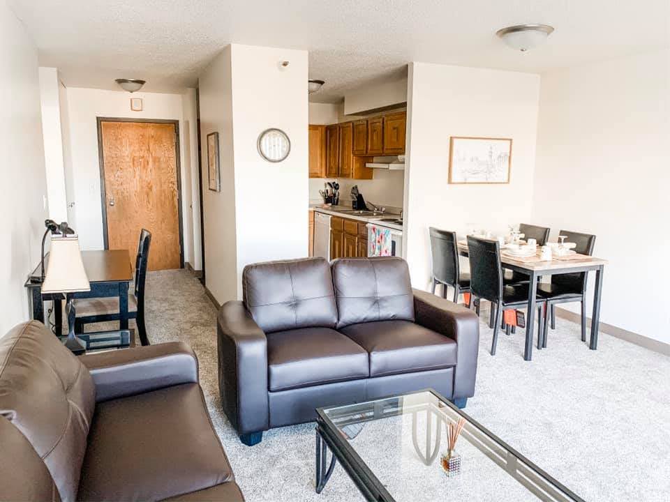 Interior view of living room example with light gray carpet, two brown leather couches glass coffee table, dining table with four chairs, adjacent to the kitchen.
