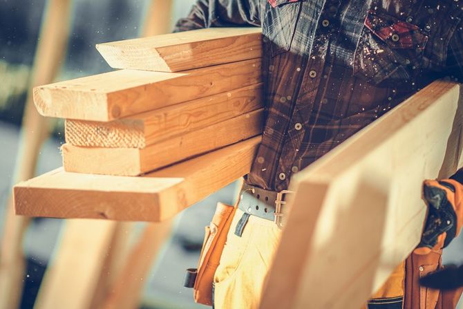 Person carrying wooden planks on a construction site, wearing a plaid shirt and tool belt.