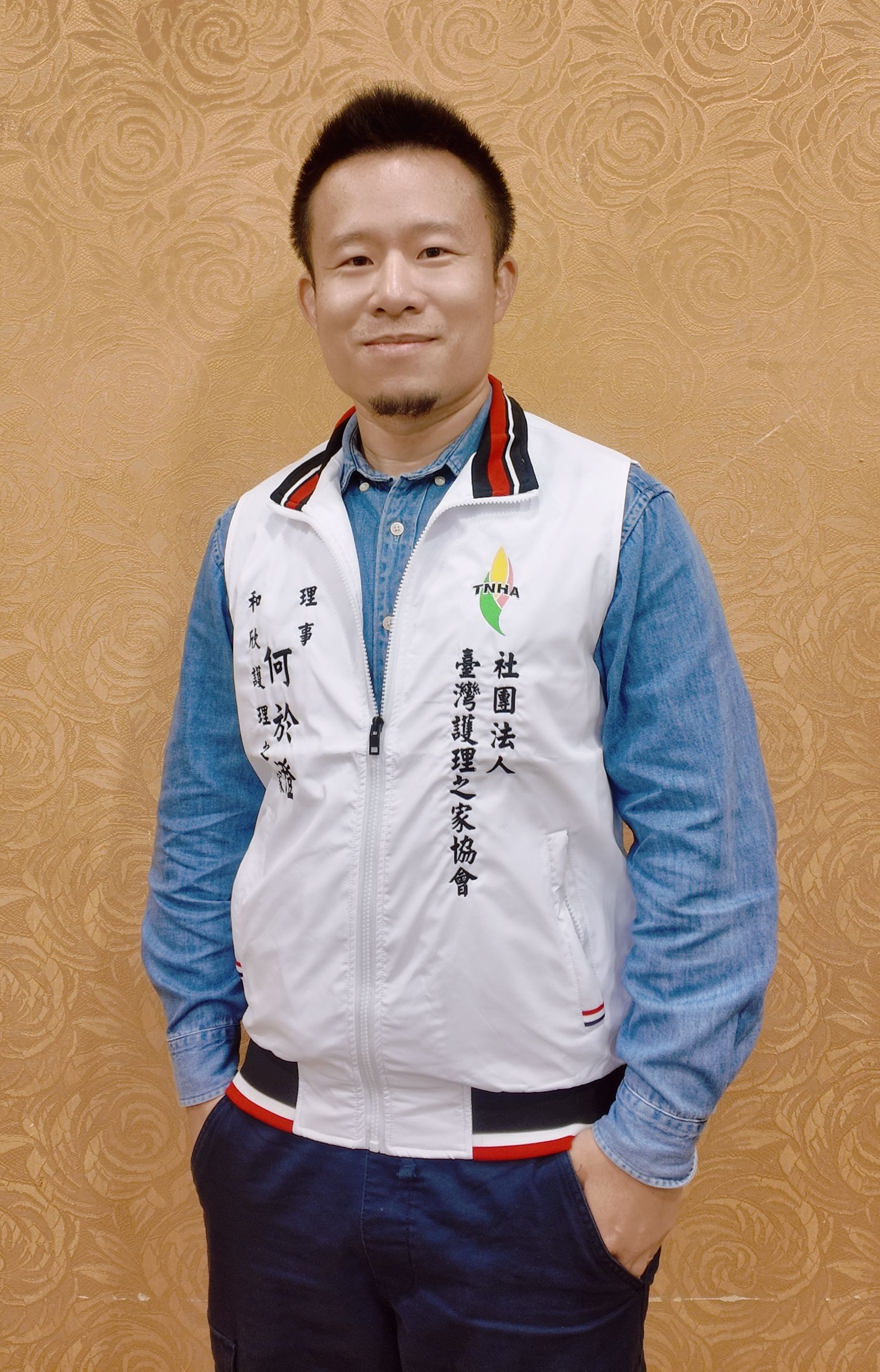 一名男子靠在棕褐色的牆上，身穿藍色襯衫，外罩白色背心，雙手插在口袋裡。