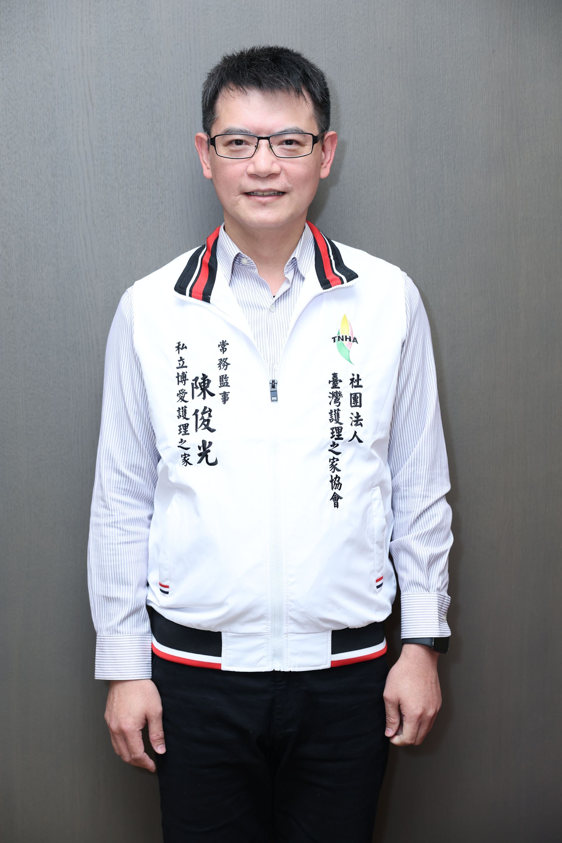 一名男子靠在灰色牆壁上，身穿白色夾克，夾克邊緣飾有黑色和紅色滾邊，下身穿著黑色褲子。