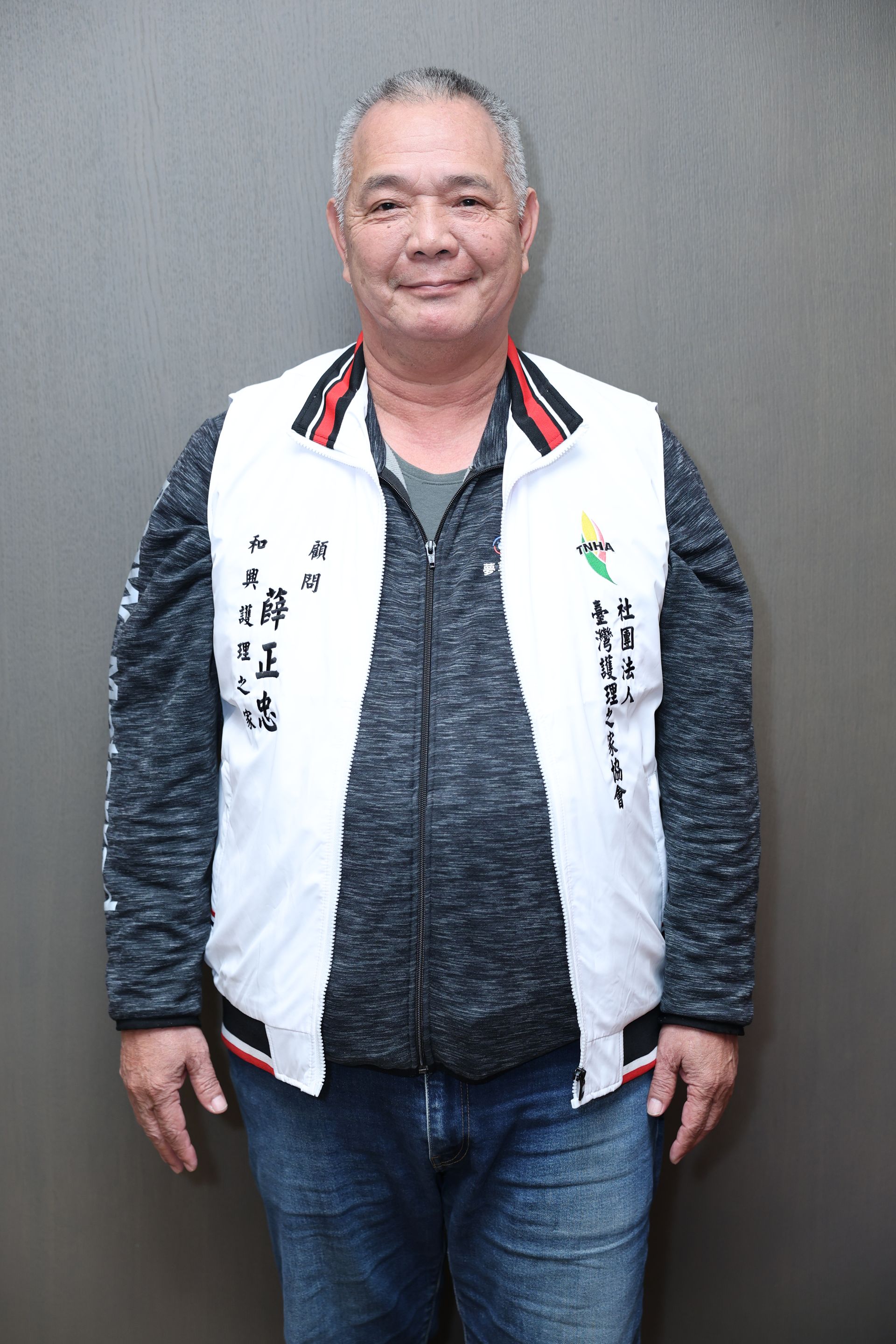 一名男子站在灰色背景前，身穿白色背心，內搭深色襯衫和牛仔褲。