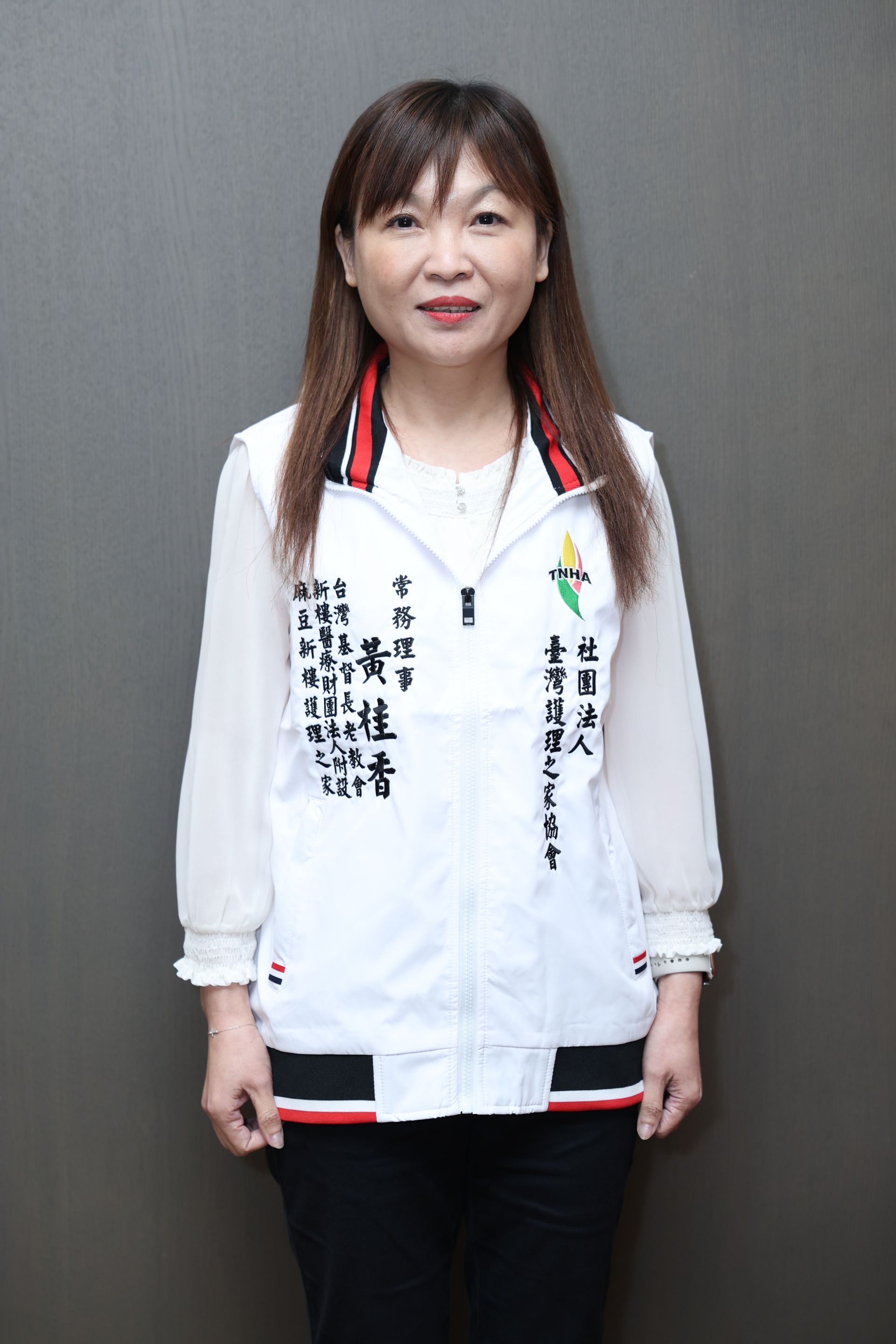 一位女子靠在灰色的牆邊，身穿白色刺繡襯衫、黑色褲子和紅色耳環。