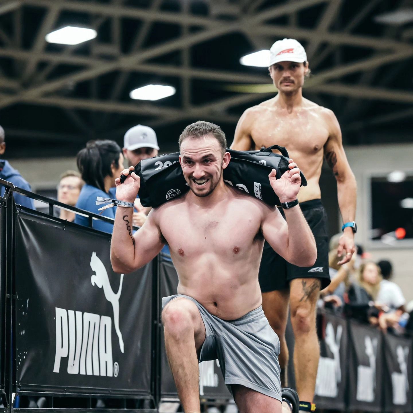 Man doing lunges with a weight bag. Another man stands behind him. Indoors with spectators and branding.