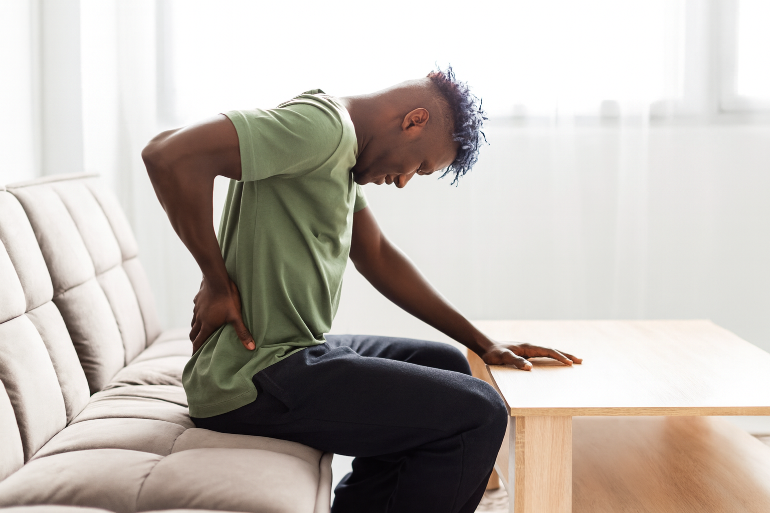 Man seated, hunched over, clutching his lower back in pain. Indoors, wearing green shirt and black pants.