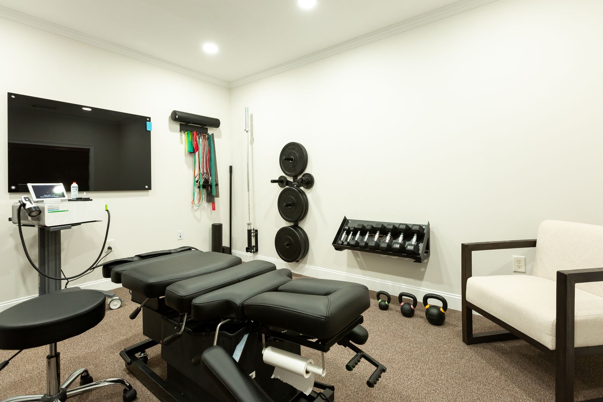 Chiropractic treatment room with equipment: table, weights, exercise bands, TV, and seating.