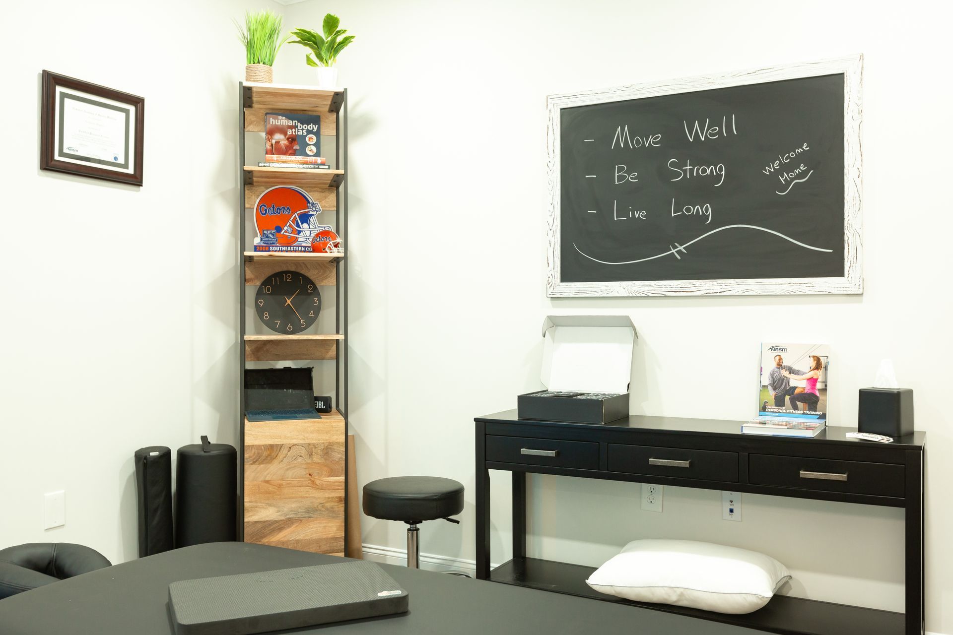 Interior of a physical therapy office. A table with equipment, desk, shelves, and a chalkboard with writing.