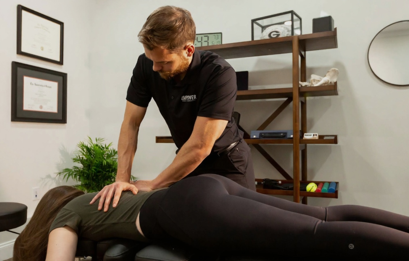 Chiropractor adjusting a patient's back in an office; man in black shirt, woman in leggings, wooden shelves.