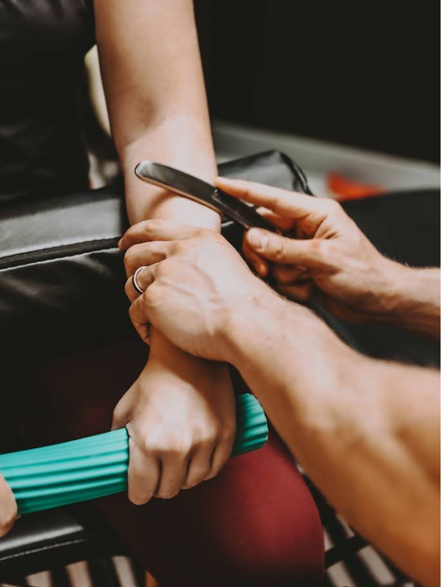 Person receiving myofascial release on their forearm with a metal tool and foam roller; indoor setting.