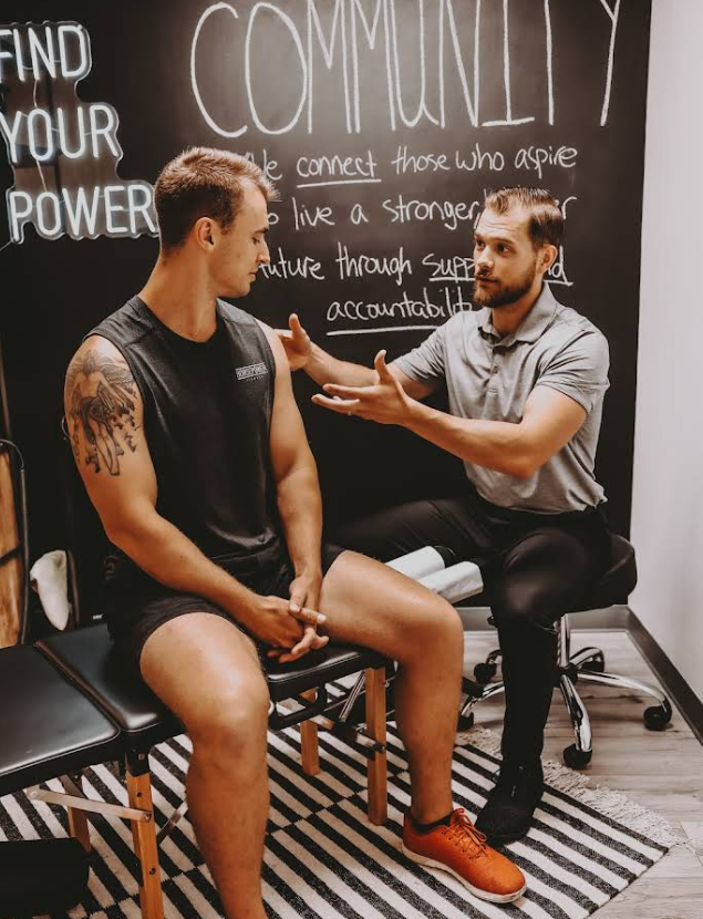 Man with shoulder tattoo consults with a doctor in a clinic. Doctor gestures, chalkboard background.