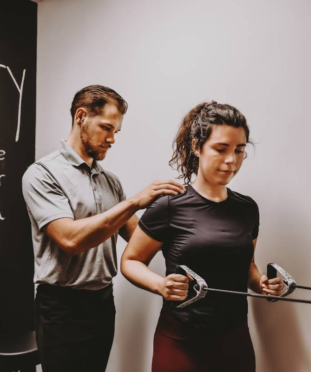 Man assisting woman with resistance band chest exercise. Indoors, white wall.