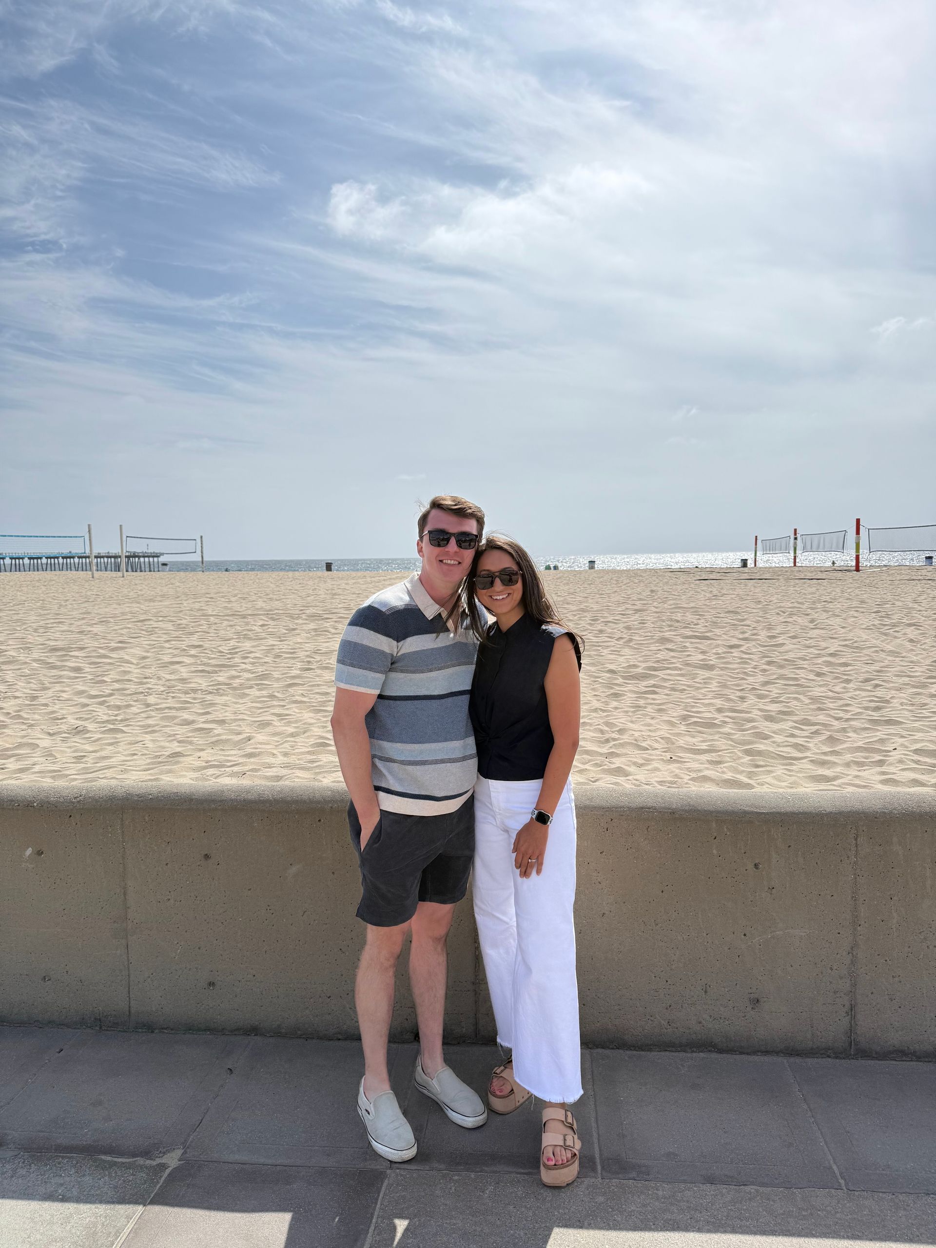 Couple on a beach: Man in striped shirt and shorts, woman in black top and white pants, posing near sand and ocean.