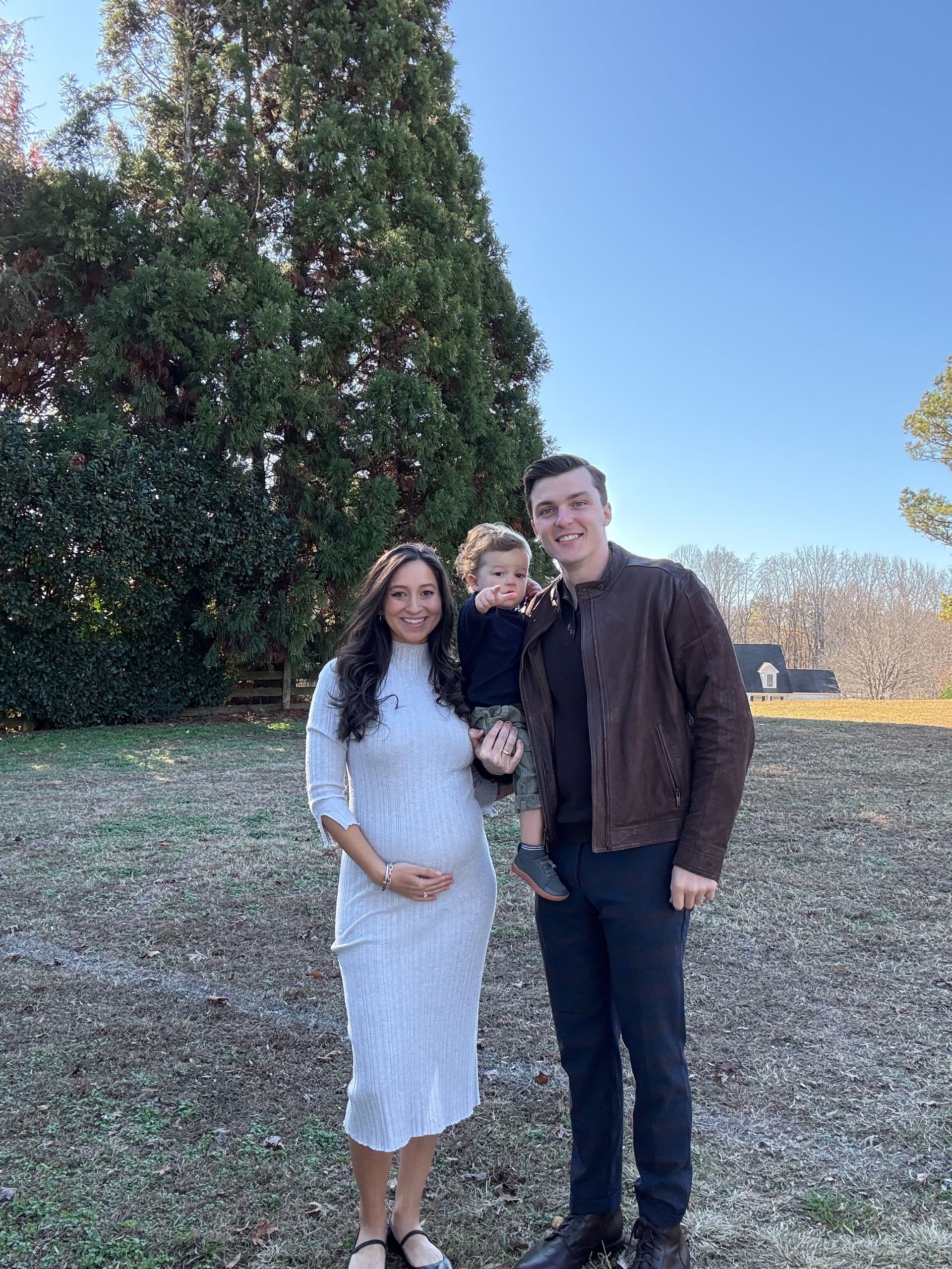 Family of three poses outdoors: pregnant woman, child in arms, man wearing a brown jacket.