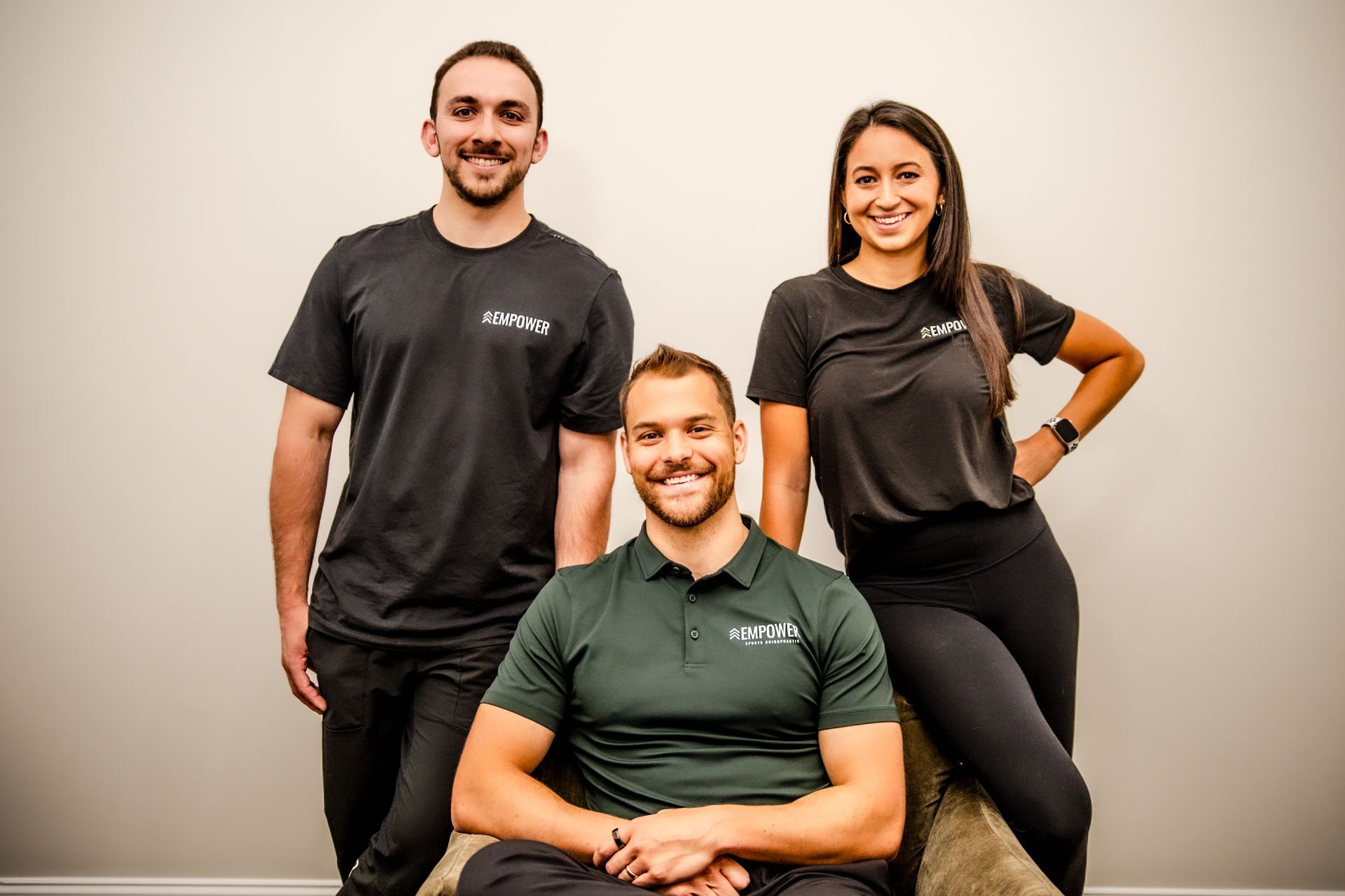 Three smiling people in athletic wear: two standing and one seated against a light wall.