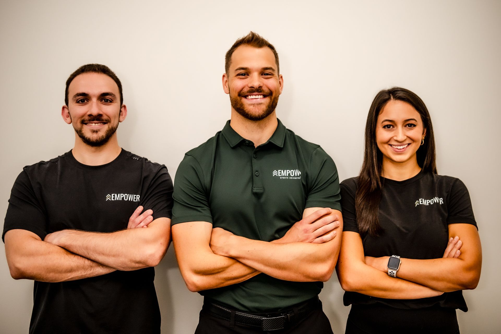 Three people with arms crossed, smiling. One in a green polo, two in black shirts in front of a neutral wall.