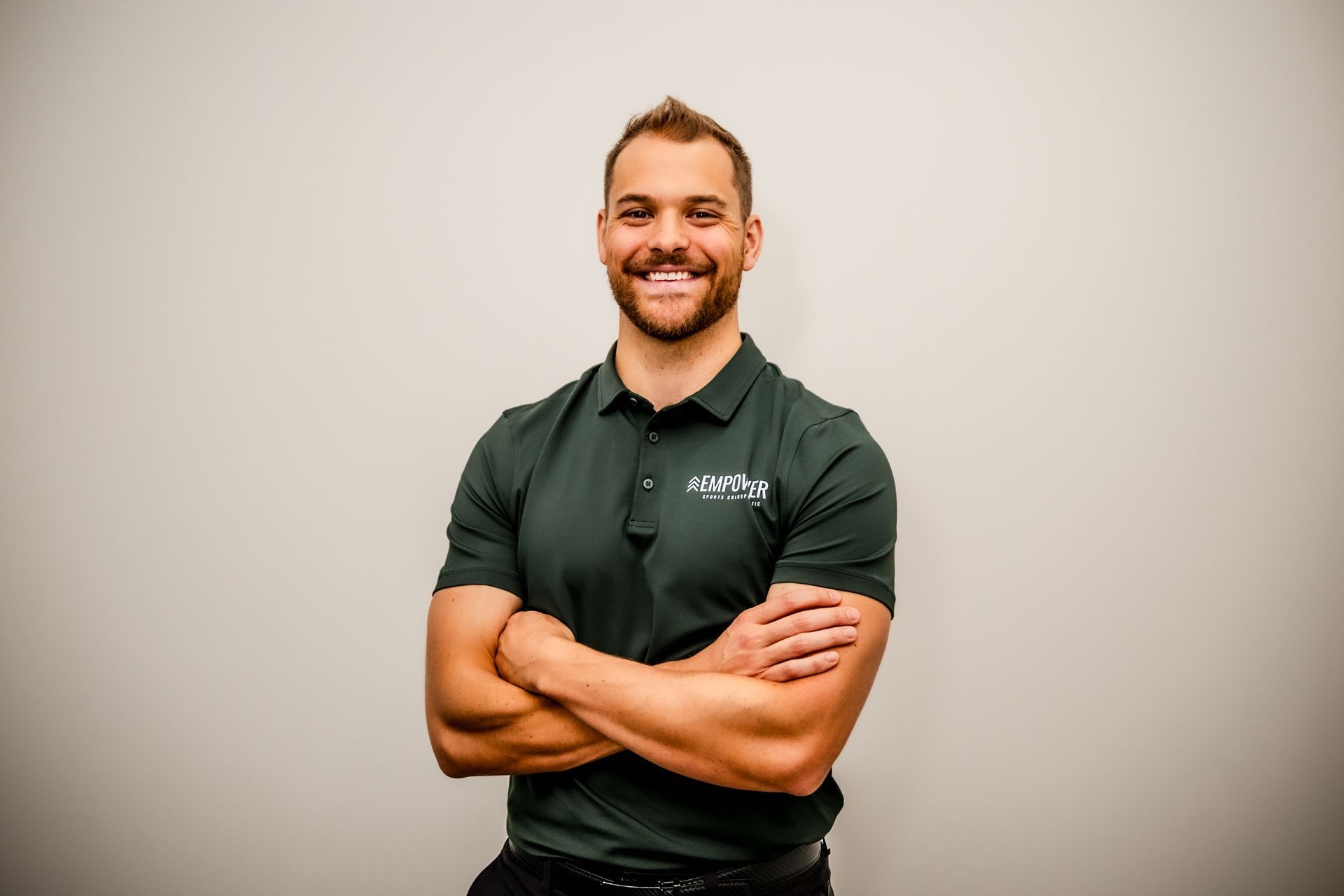Man in green polo shirt, arms crossed, smiling, standing in front of a neutral wall.