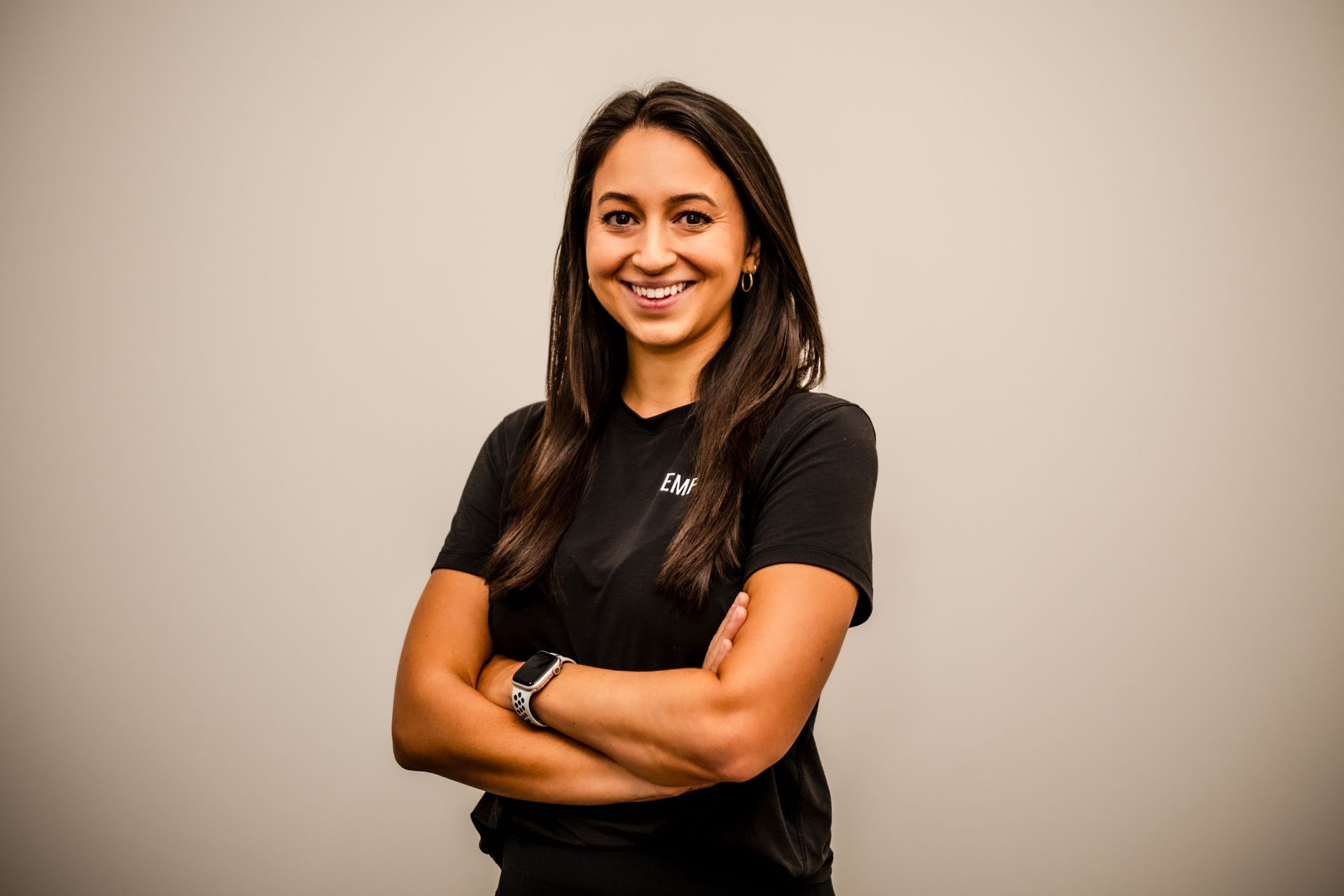 Woman with arms crossed, smiling, wearing a black t-shirt and smartwatch, standing against a neutral background.