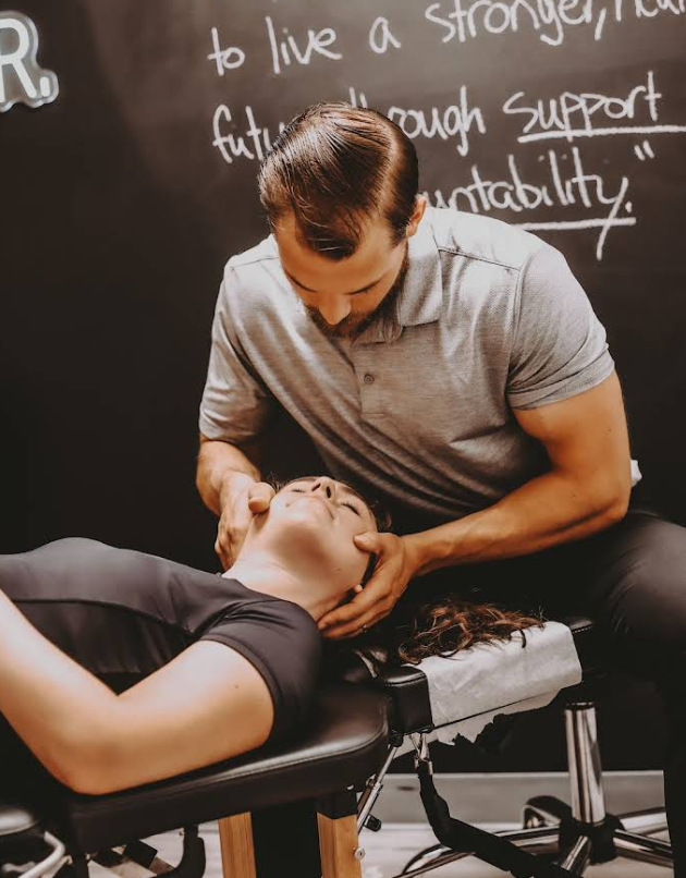 Man adjusting a woman's neck at a chiropractic office. They're indoors, the background is a chalkboard with text.