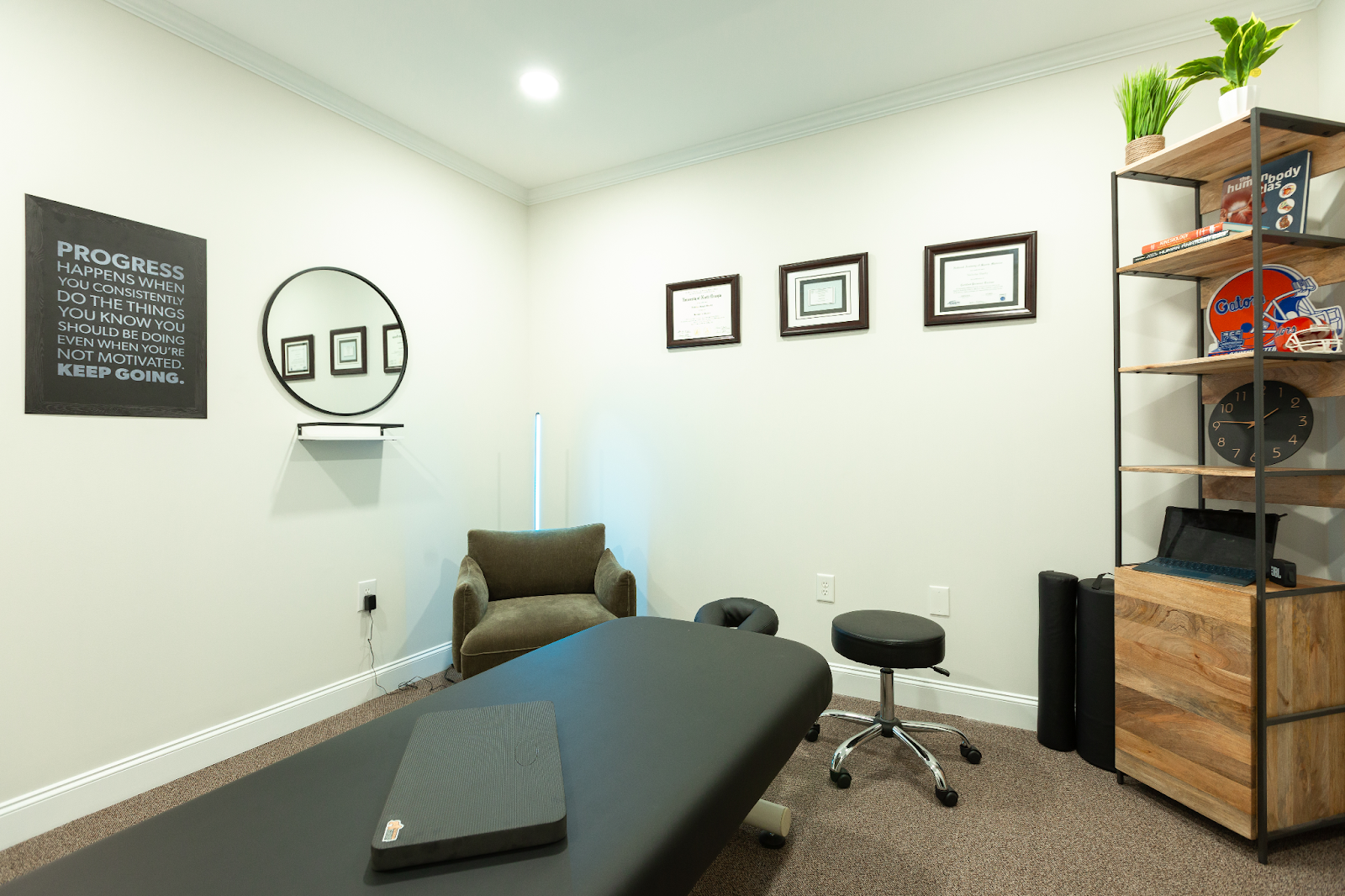 Physiotherapy room with a massage table, chair, stool, and shelving unit. White walls and neutral tones.