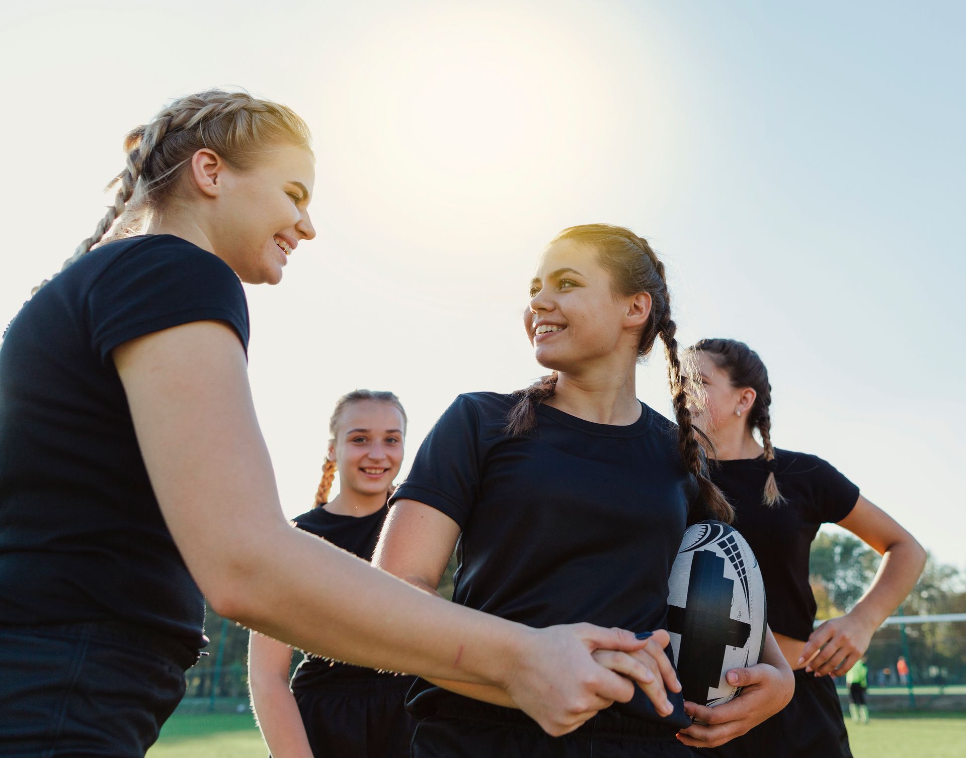 Female rugby players smiling and shaking hands on a sunny field, one holding a rugby ball.