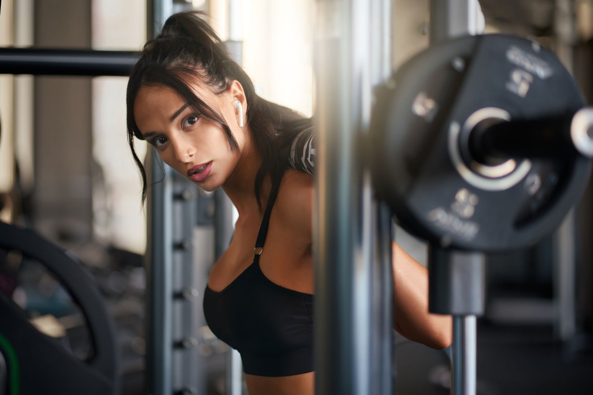 Woman in black sports bra at the gym, looking over her shoulder. Barbell in the foreground.