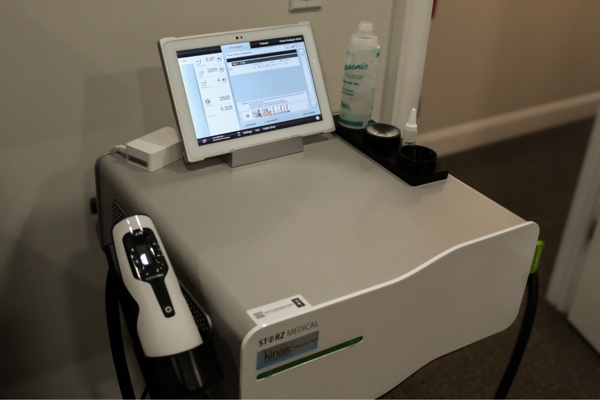White medical device with touchscreen and handheld wand in a hallway, with a bottle on top.