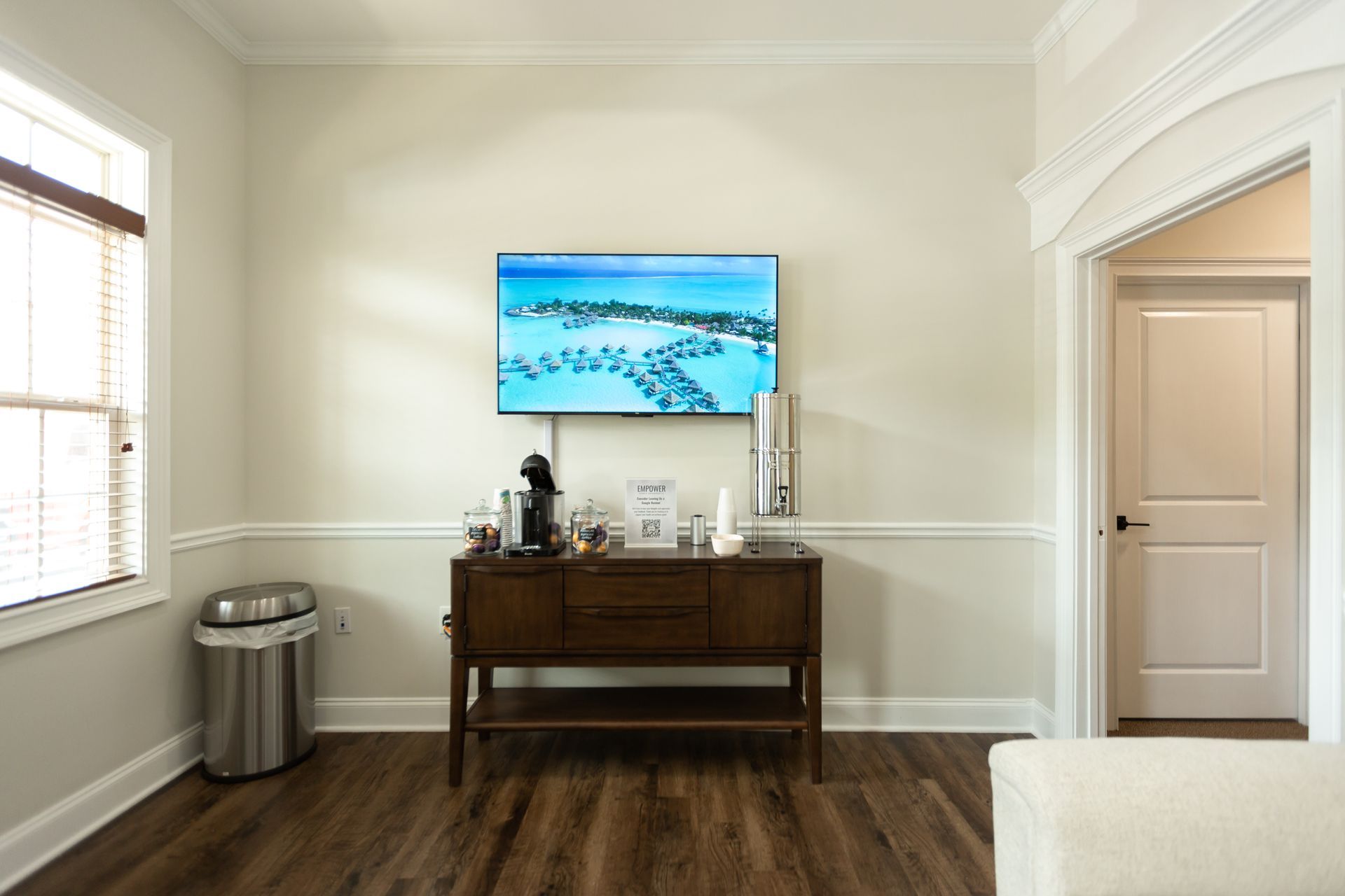 Room with a mounted TV, wood cabinet, window, door, and a trash can on a hardwood floor.