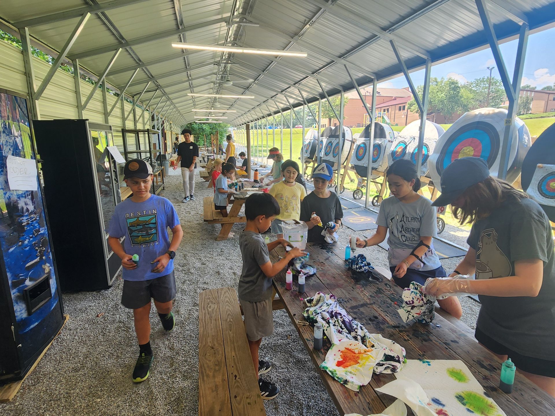 People arranging archery crafts at a long table under a pavilion, with a blue vending machine and colorful display in back