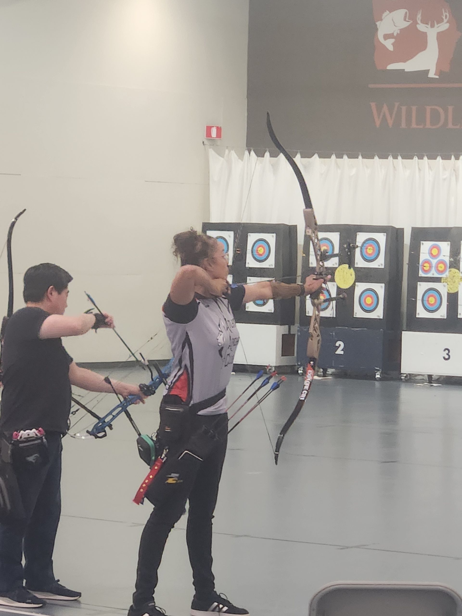 Two archers aiming bows at targets indoors during a competition