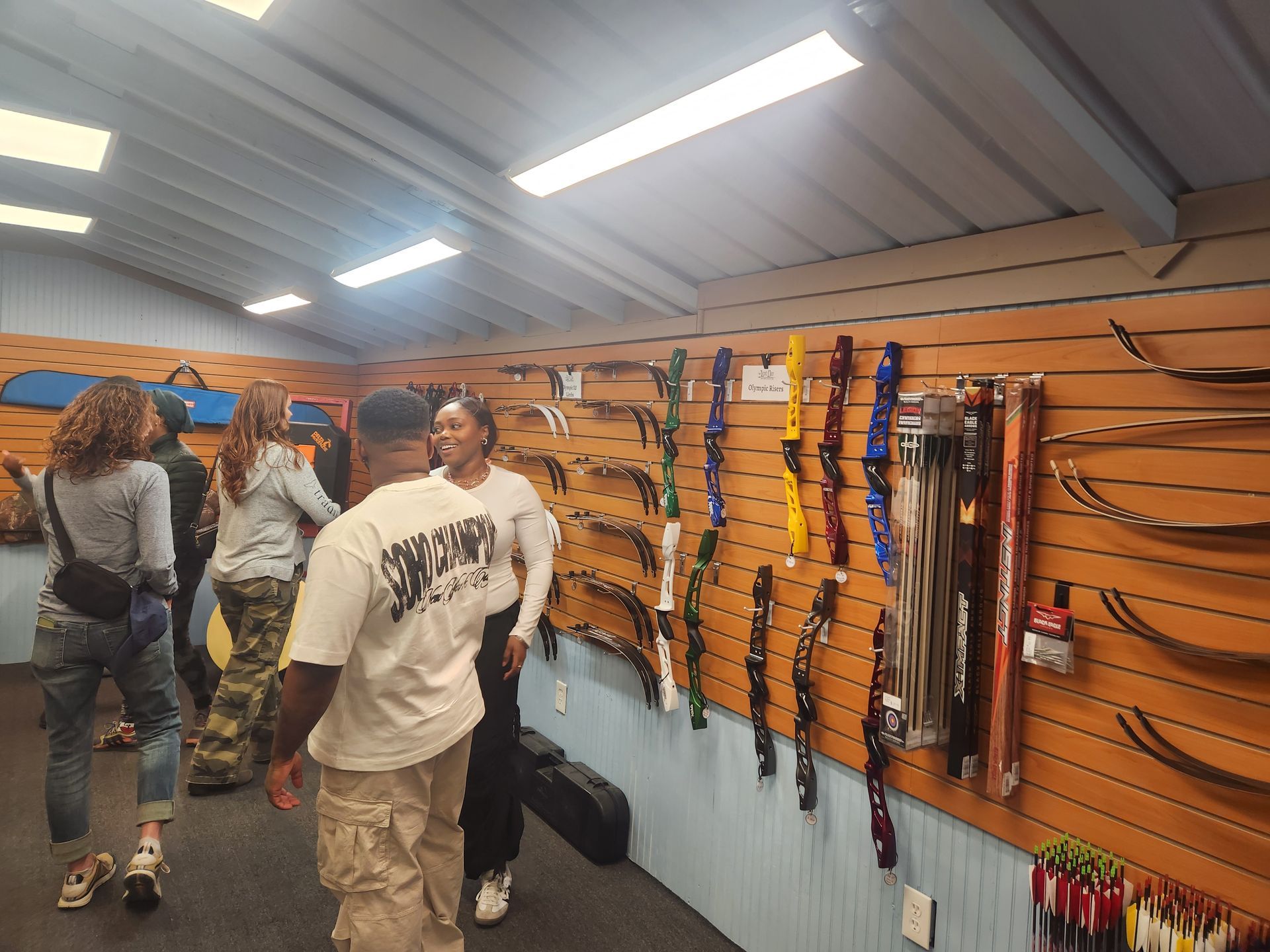 People browsing archer rods in a tackle shop with wood-paneled walls and bright overhead lights