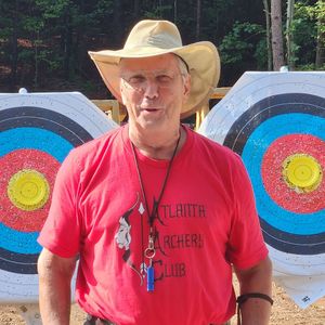 Man wearing a red Atlanta Archery Club shirt, sunglasses, and a hat stands in front of archery targets.