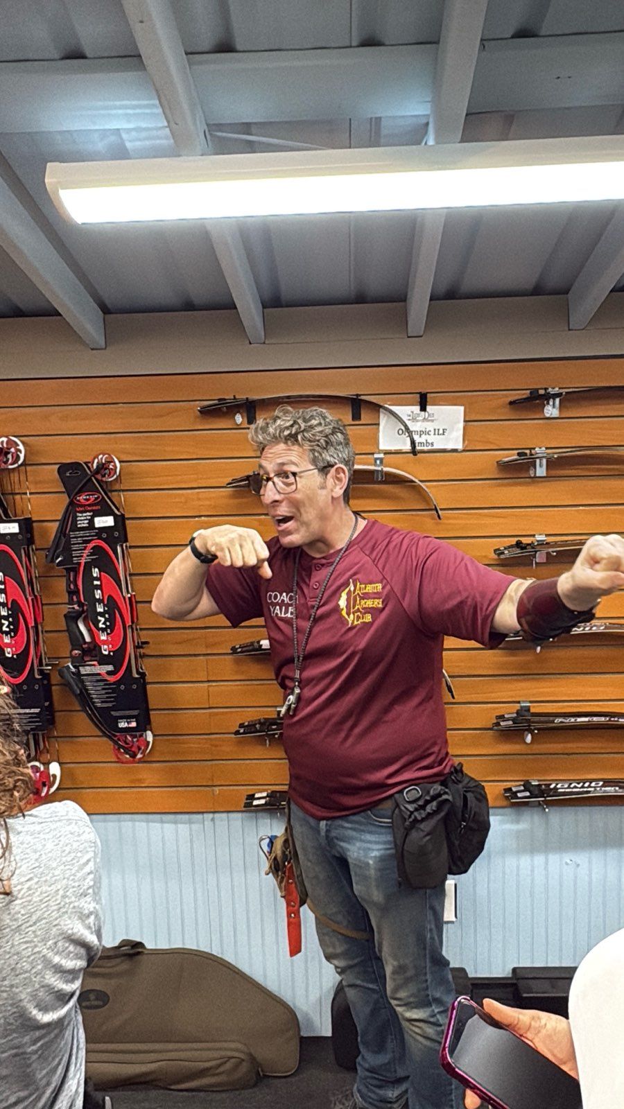 Man in maroon shirt gesturing inside a gear shop with hanging bags and wall-mounted racks