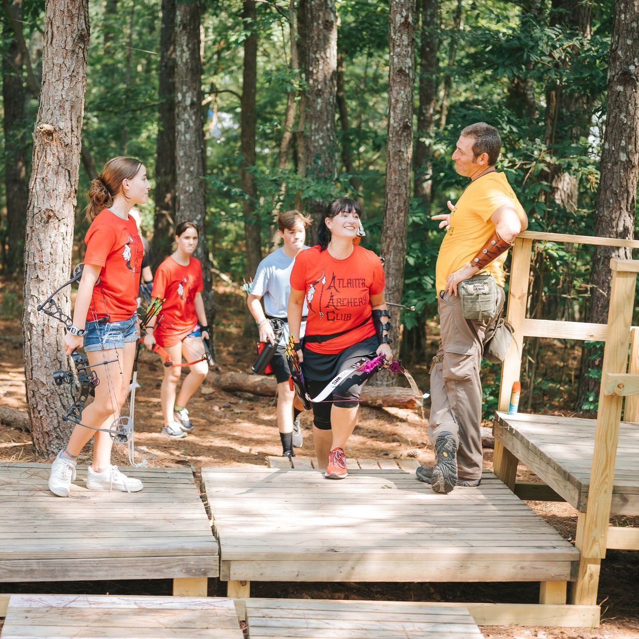 Group of people on wooden platforms in a forest, being instructed by a person in orange.