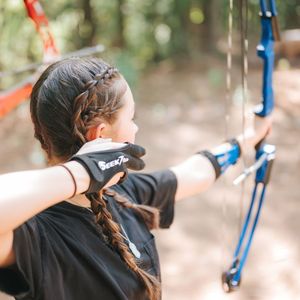 Person with braided hair aiming a blue bow and arrow outdoors.