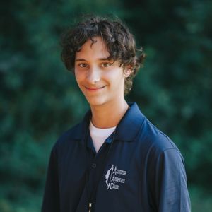 Young person with dark curly hair smiling in a navy polo shirt with logo, in front of green foliage.