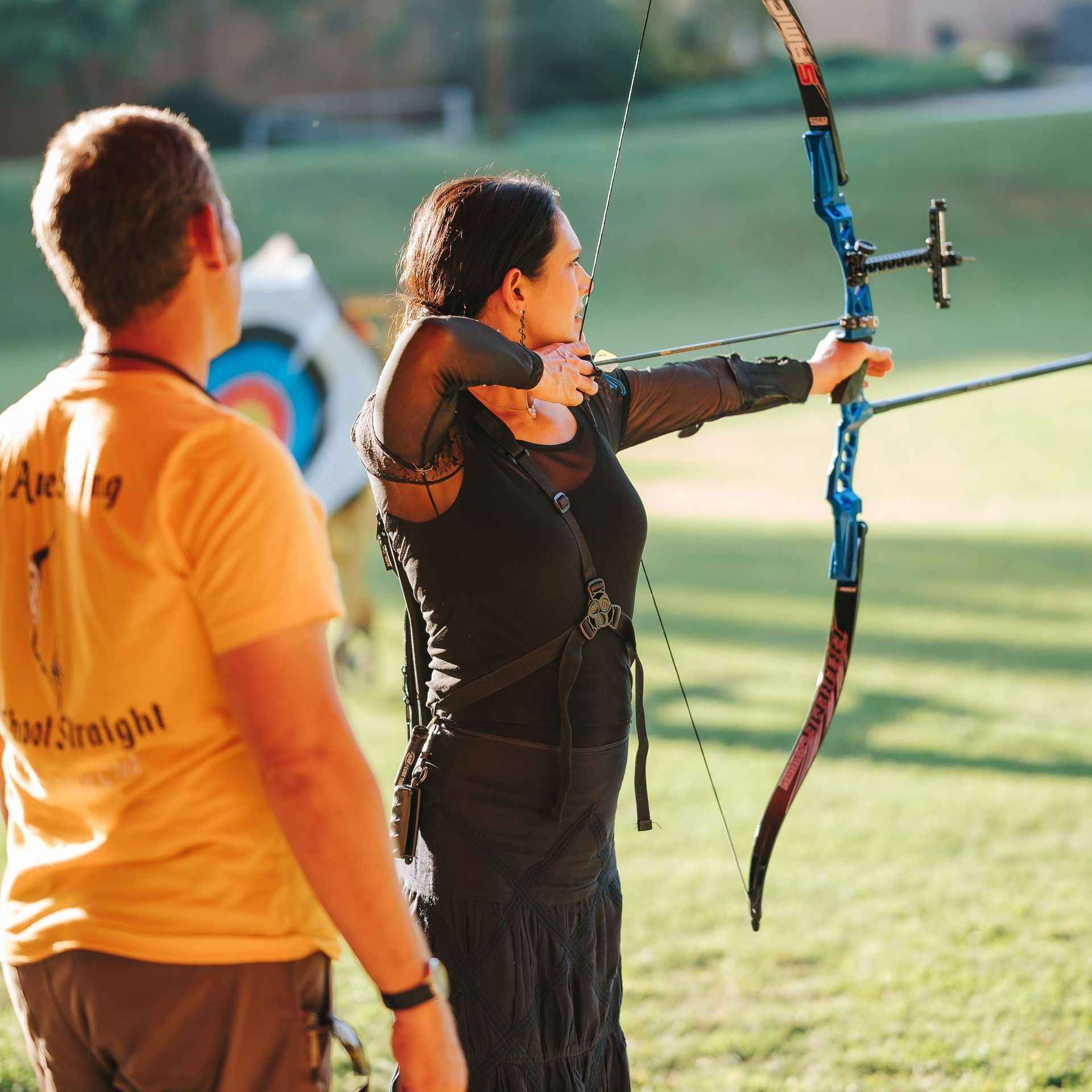 Woman aiming a blue bow at a target, with a man watching. Green field and sunny outdoor setting.