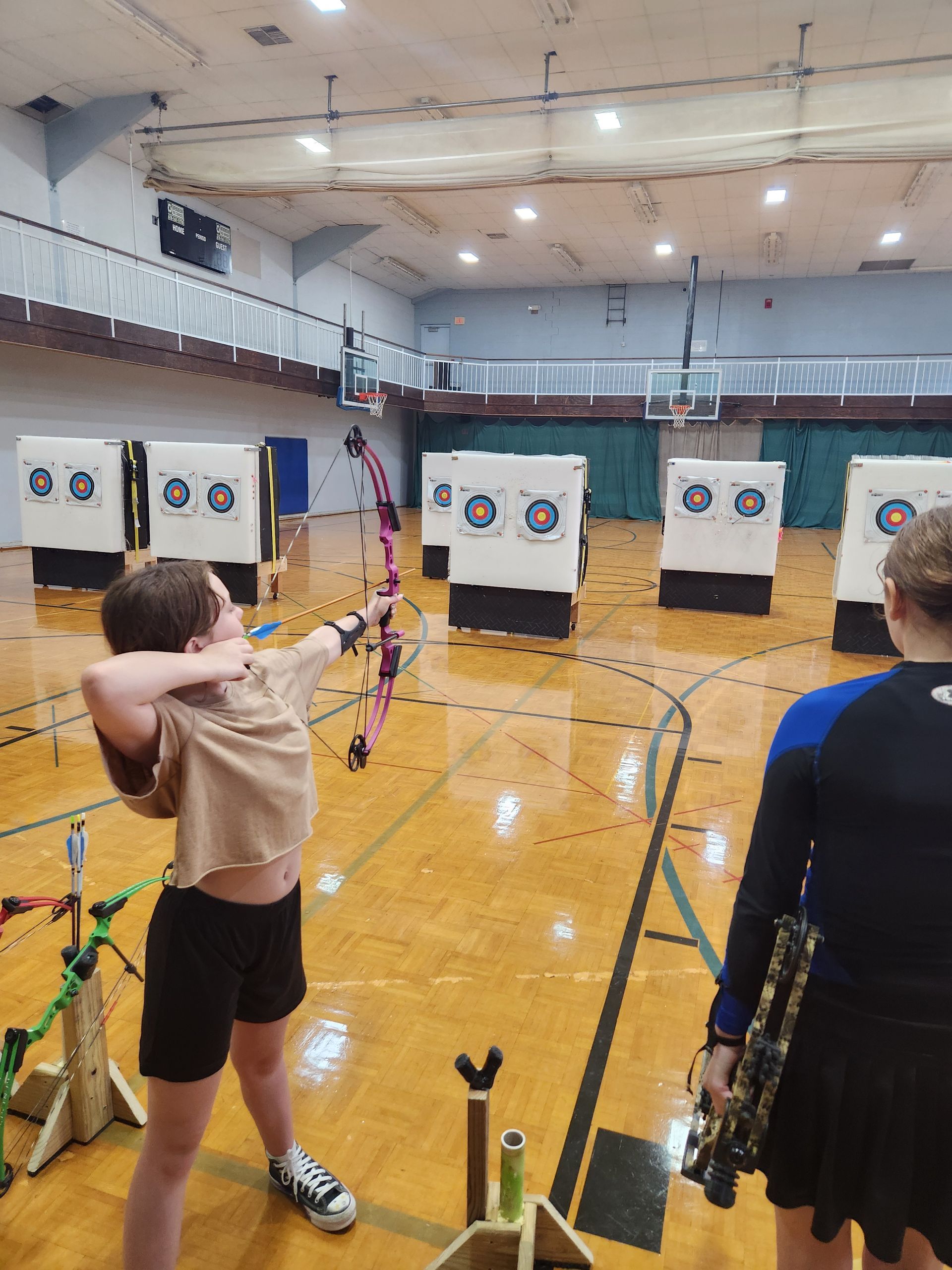 Two archers aiming at targets in a gym with multiple archery targets lined up ahead