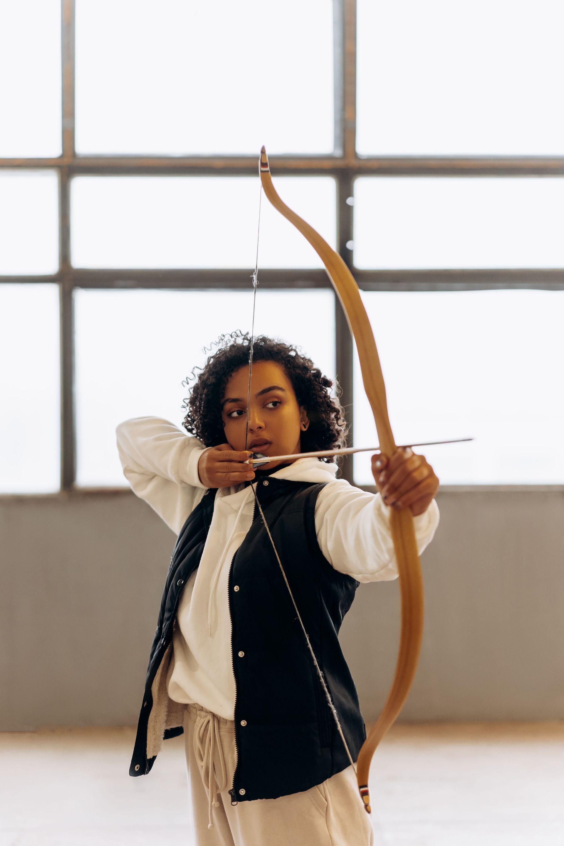 Person drawing a bow indoors, aiming an arrow in front of a large window.