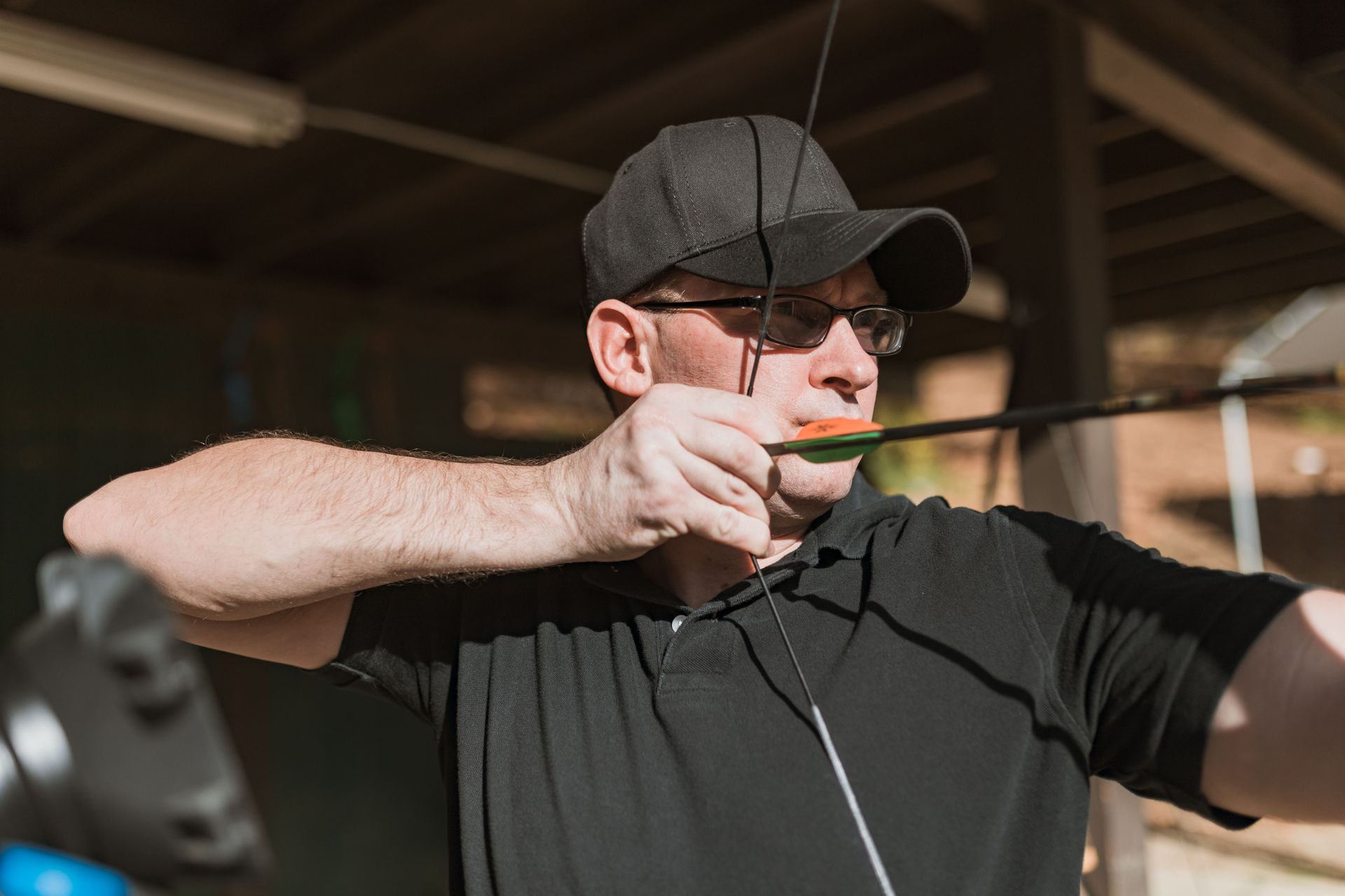 Man in a black cap aiming a bow and arrow outdoors under a covered range