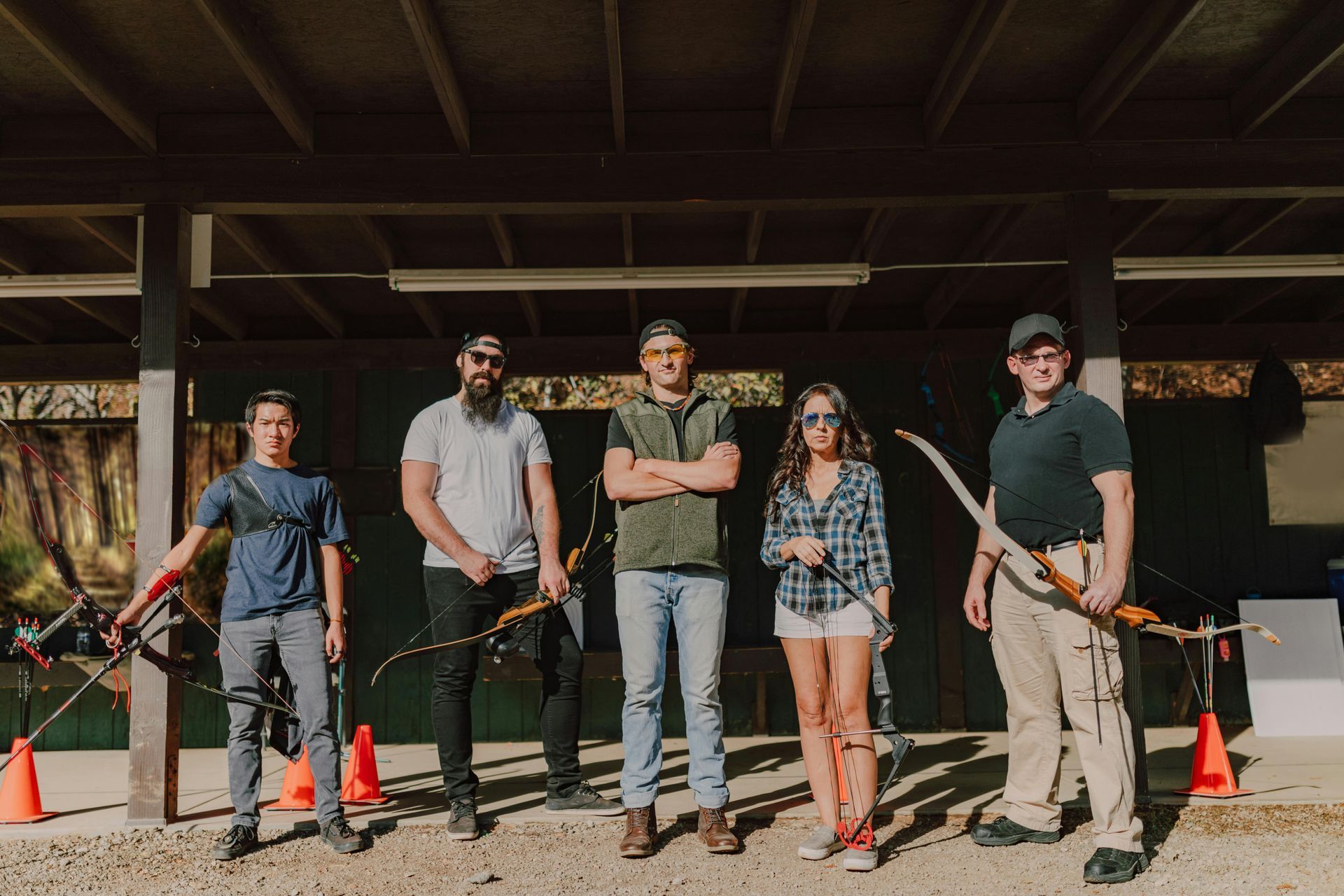 Five people standing with rifles under a wooden shelter, with orange cones and target frames behind them.