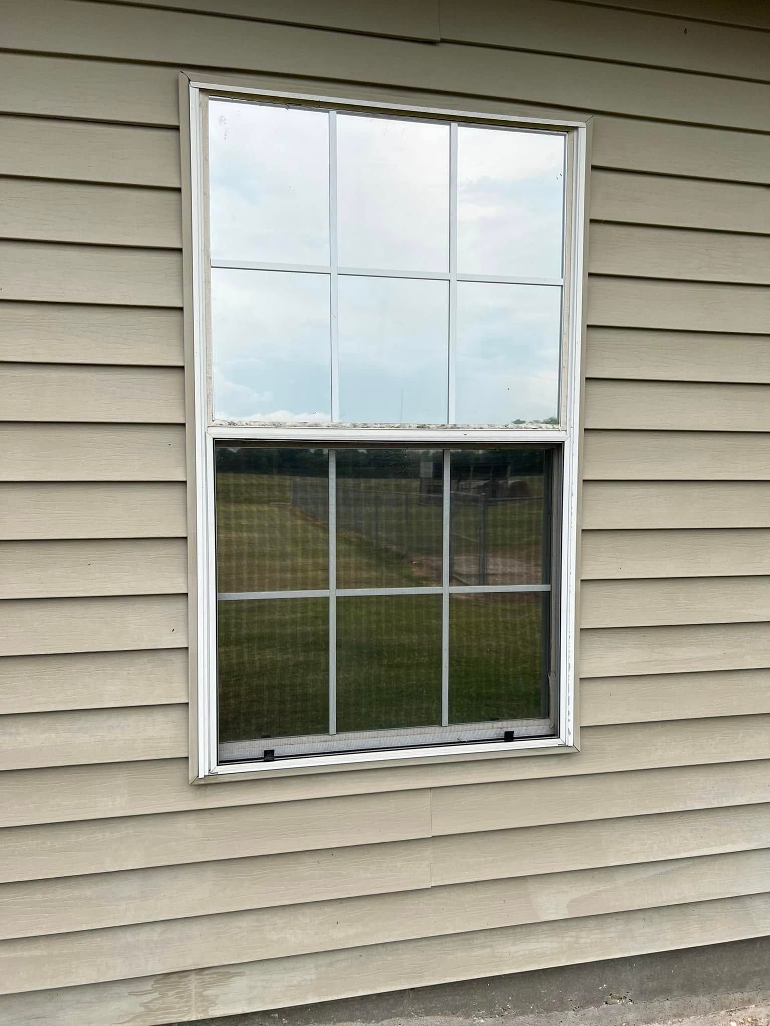 White-framed window on beige siding with two glass panes, reflecting sky and showing a grassy yard.
