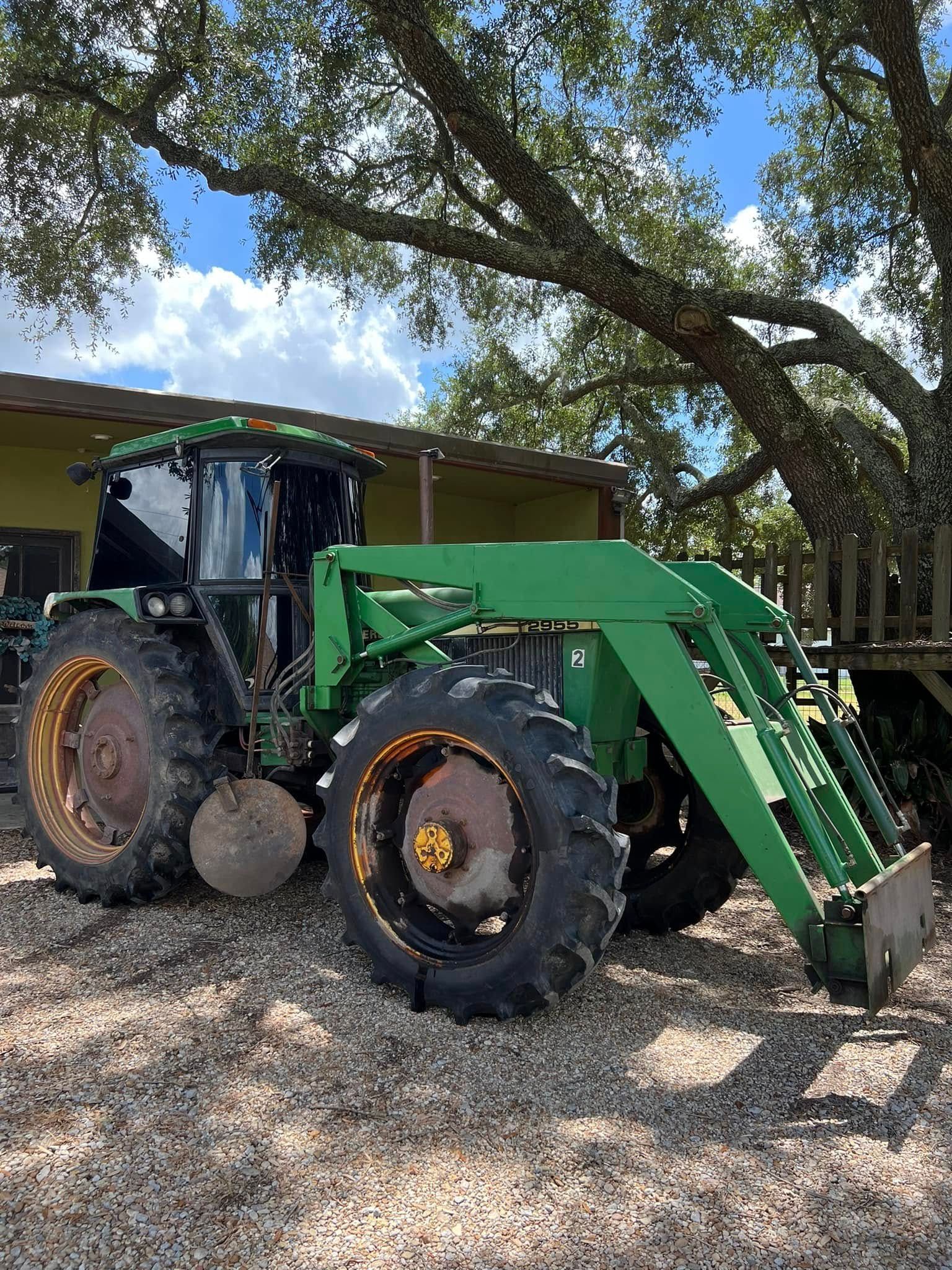 Green tractor with front loader parked on gravel under trees beside a building