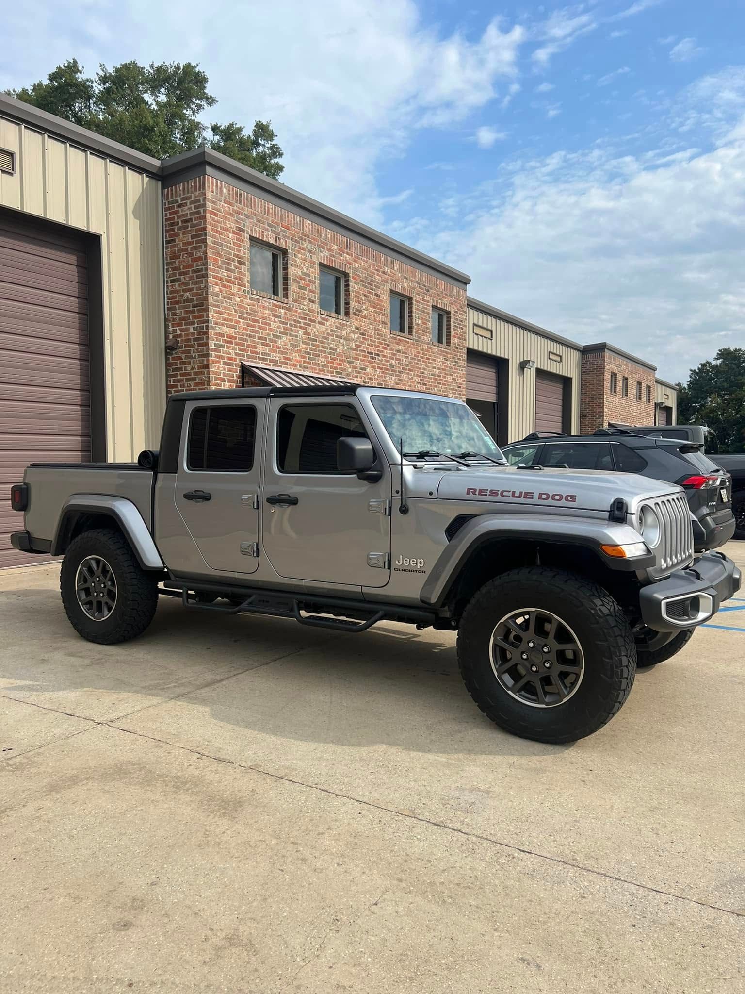 Gray lifted Jeep Gladiator parked outside brick industrial buildings under a blue sky.